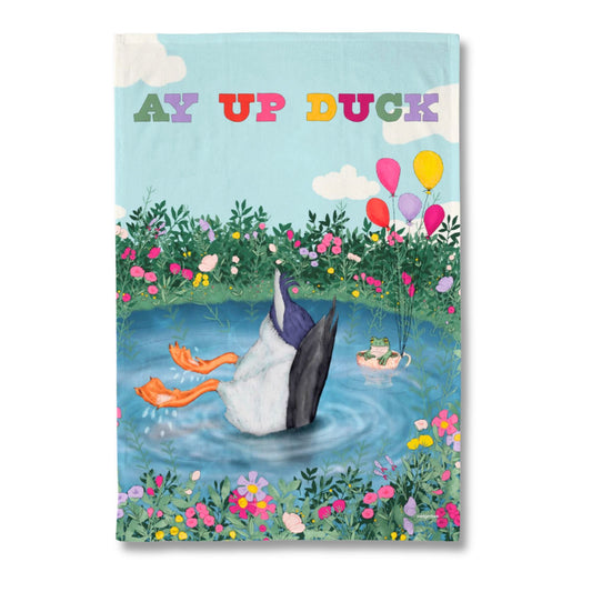 A rectangular printed tea towel shown against a white background. The design features a whimsical illustration of a duck standing in a pond with its legs in the air, surrounded by flowers, balloons, and the text 'AY UP DUCK' at the top.