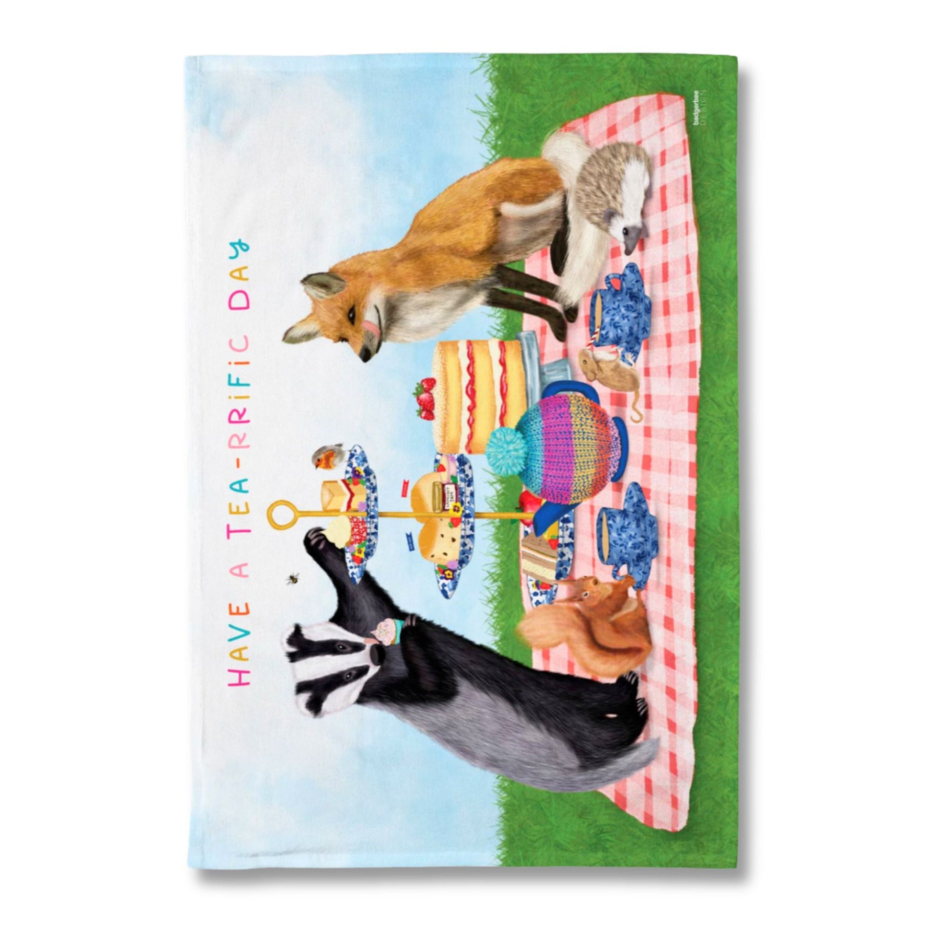 A rectangular printed tea towel featuring a whimsical outdoor picnic scene with a fox, a raccoon, a fox toy, teacups, cakes, and the text 'HAVE A TEA-RRIFIC DAY' on a white background.