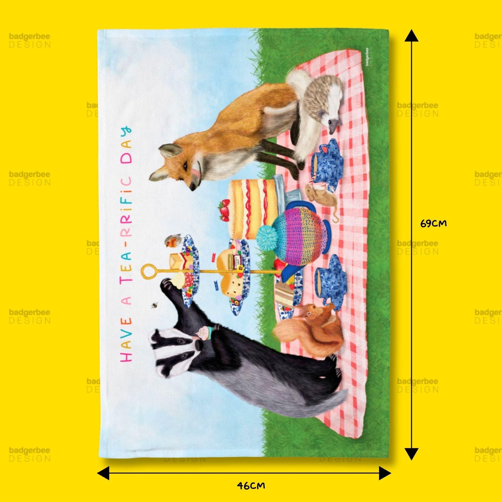 A rectangular printed tea towel shown against a bright yellow background, with dimension arrows and labels indicating 69 cm height and 46 cm width. The printed design features a picnic scene with a fox, a black-and-white cat, a striped blanket, flowers, and the text 'HAVE A TEARFFIC DAY'.