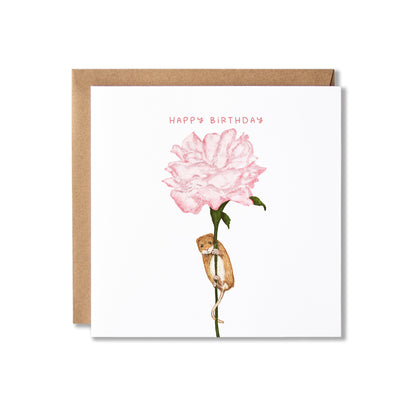 A square white birthday card with a kraft brown envelope behind it, featuring an illustration of a pink flower with a small brown mouse hanging from the stem and the words 'HAPPY BIRTHDAY' in pink at the top.