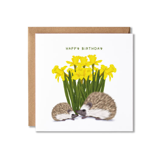 A square birthday card with a brown envelope, featuring an illustration of two hedgehogs, one big, one small, facing a bouquet of yellow daffodils, with the text 'HAPPY BIRTHDAY' above.