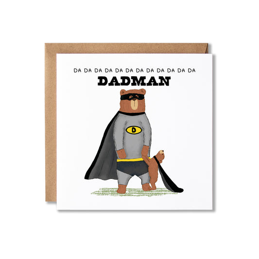 A square white greeting card with a brown kraft envelope behind it, featuring an illustration of a superhero bear in a gray cape and mask, holding hands with a bear cub and the word 'DADMAN' above, on a light gray background.