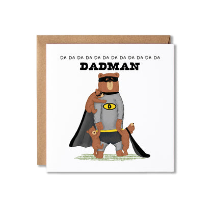 A square white greeting card with a kraft brown envelope behind it, featuring an illustration of a bear dressed as a superhero and three bear cubs hanging on to him, with the word 'DADMAN' and repeated 'DA' text above. The card is photographed on a clean white background.