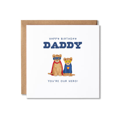 A square birthday card with a kraft envelope behind it, featuring the text 'HAPPY BIRTHDAY DADDY' and 'YOU'RE OUR HERO!' with two illustrated super-hero lion cubs on a white card background.