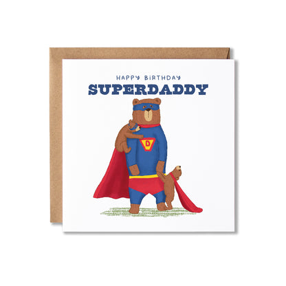A square birthday card with a kraft envelope behind it, featuring an illustration of a superhero bear with a bear cub on his shoulder and holding hands with another bear cub. The card reads 'HAPPY BIRTHDAY SUPERDADDY' in blue text on a white background.