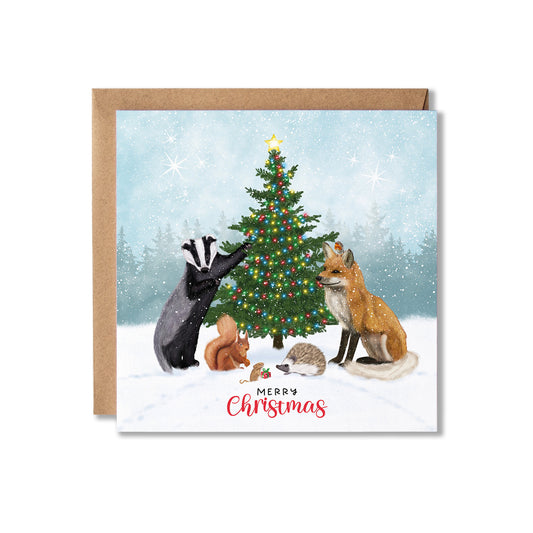 A square Christmas greeting card with a kraft envelope, featuring an illustrated winter scene with a decorated tree, a badger, a fox, and small woodland animals in the snow. The card reads 'Merry Christmas' at the bottom.