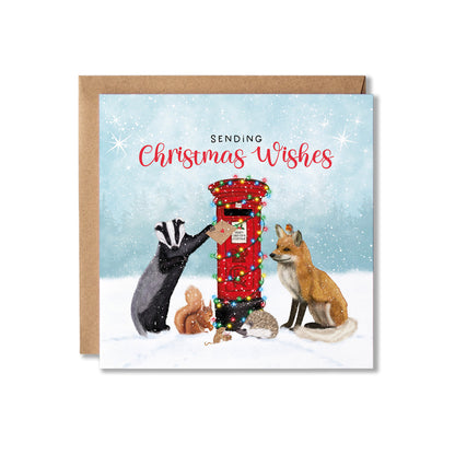 A square Christmas greeting card with a kraft envelope behind it. The card illustration shows woodland animals around a red post box decorated with colorful lights, with the text 'SENDING Christmas Wishes' on a snowy blue background.