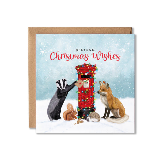 A square Christmas greeting card with a kraft envelope behind it. The card illustration shows woodland animals around a red post box decorated with colorful lights, with the text 'SENDING Christmas Wishes' on a snowy blue background.