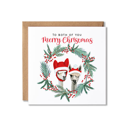 A square white Christmas greeting card with a kraft envelope behind it. The card reads 'TO BOTH OF YOU Merry Christmas' above an illustration of two alpacas wearing red Santa hats, framed by a green pine wreath with red berries, on a white background.