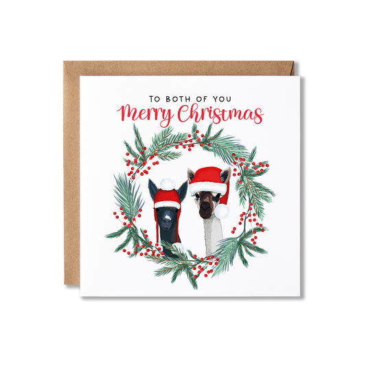 A square Christmas greeting card with a kraft envelope behind it. The card reads 'TO BOTH OF YOU Merry Christmas' above an illustration of two alpacas wearing Santa hats inside a green pine wreath with red berries on a white background.
