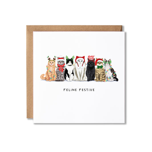 A square greeting card with a brown kraft envelope behind it, featuring an illustration of seven cats wearing Christmas hats and scarves, with the text 'FELINE FESTIVE' below them, on a white background.