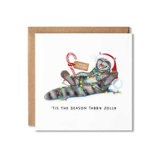 A square white greeting card with a brown kraft envelope behind it, featuring an illustration of a gray tabby cat wrapped in Christmas lights and a red Santa hat, with the text “'Tis the season tabby jolly” below.