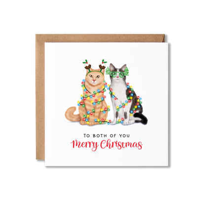 A square Christmas greeting card with a kraft envelope, featuring an illustration of two cats wrapped in colorful holiday lights and wearing green glasses and reindeer antlers, with the text 'TO BOTH OF YOU Merry Christmas' on a white card background.