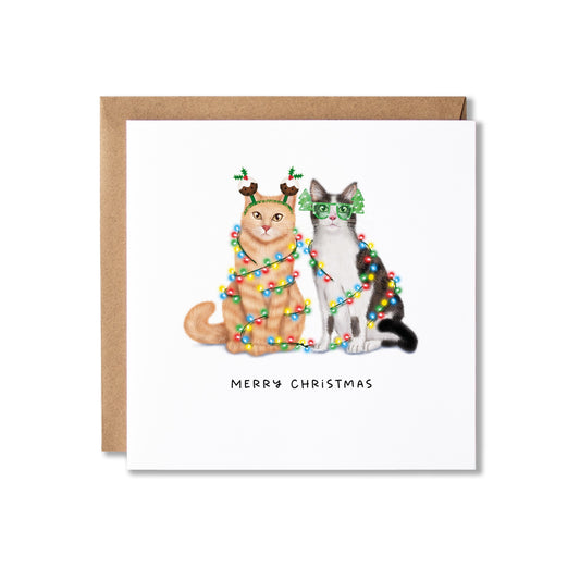 A square Christmas greeting card with a brown kraft envelope, featuring an illustration of two cats wrapped in colorful holiday lights and wearing festive accessories, with the text 'MERRY CHRISTMAS' below.