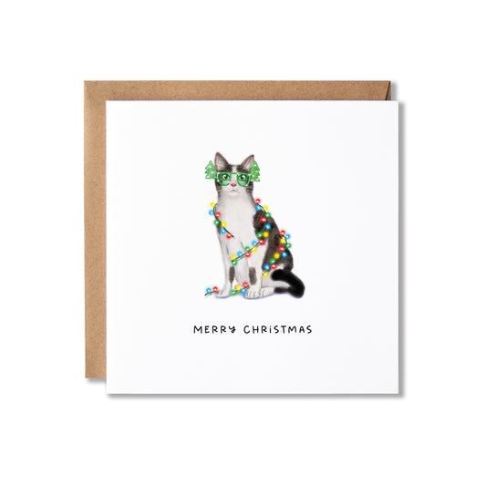 A square Christmas greeting card with a brown kraft envelope behind it. The card features an illustration of a black and white cat wearing green Christmas tree glasses, with the text 'MERRY CHRISTMAS' printed below.
