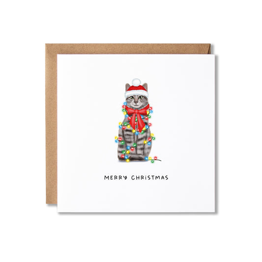 A square white Christmas greeting card with a brown kraft envelope behind it. The card features an illustration of a gray tabby cat wearing a red Santa hat and red bow tie, wrapped in multicolored Christmas lights, with the text 'MERRY CHRISTMAS' below.
