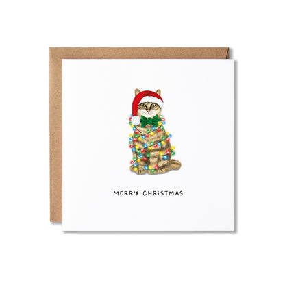 A square white Christmas greeting card with a brown kraft envelope behind it. The card features an illustration of a tabby cat wearing a red Santa hat and green bow tie, wrapped in multicolored Christmas lights, with the text 'MERRY CHRISTMAS' below.