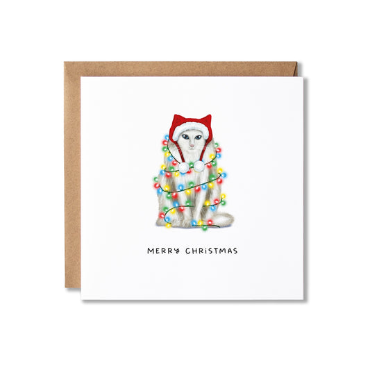 A square Christmas greeting card with a white front, featuring an illustration of a white cat wearing a red Santa hat and wrapped in multicolored holiday lights. The card is shown with a brown kraft envelope behind it on a white background.