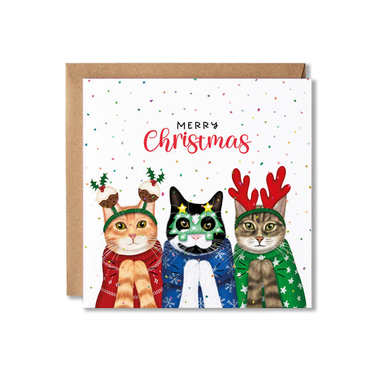 A square Christmas greeting card with a kraft envelope behind it. The card features three illustrated cats wearing holiday sweaters, scarves, and reindeer antlers, with the text 'Merry Christmas' in red script on a white background with scattered confetti.