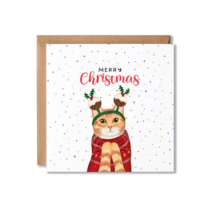 A square Christmas greeting card with a brown kraft envelope behind it. The card has a white background with scattered confetti dots, the words 'MERRY Christmas' in red and black, and an illustration of an orange tabby cat wearing a green reindeer headband and red sweater.