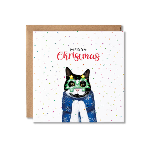 A square Christmas greeting card with a brown kraft envelope behind it. The card features a watercolor-style black-and-white cat wearing green Christmas glasses and a blue sweater, with the words 'MERRY Christmas' above it and small multicolored confetti scattered across a white background.