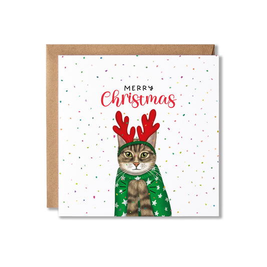 A square Christmas greeting card with a brown kraft envelope behind it. The card has a white background with scattered confetti dots and the words “Merry Christmas” above an illustration of a tabby cat wearing red reindeer antlers and a green holiday scarf with white stars.