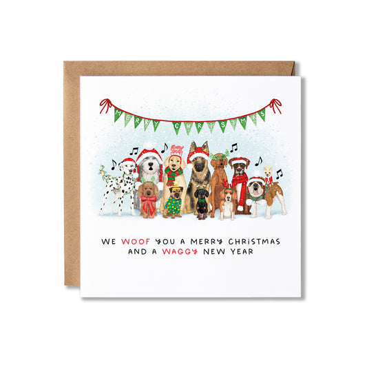 A square Christmas greeting card with a kraft envelope behind it, featuring an illustration of various dog breeds wearing Santa hats and scarves under a green bunting banner. The text at the bottom reads, 'WE WOOF YOU A MERRY CHRISTMAS AND A WAGGY NEW YEAR'.