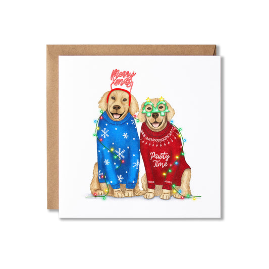 A square greeting card with a brown envelope behind it, featuring an illustration of two dogs wearing holiday sweaters and colorful lights. The left dog wears a blue sweater with white snowflakes and a red 'Merry Christmas' headband.