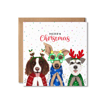 A square Christmas greeting card with a kraft envelope behind it, featuring three illustrated dogs wearing festive holiday accessories and sweaters, with the text 'Merry Christmas' on a white background with confetti.