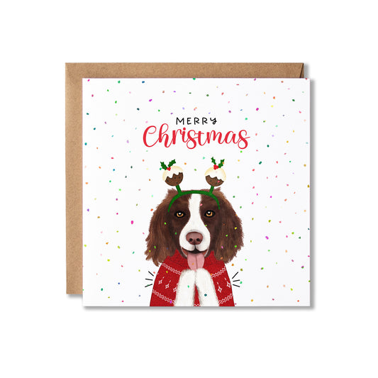 A square Christmas greeting card with a brown and white dog wearing a red sweater and Christmas pudding bobblers, with the words 'Merry Christmas' above. The card is shown with a brown envelope behind it on a white background.