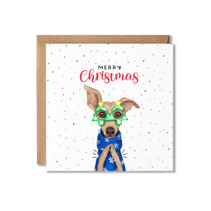 A square Christmas greeting card with a brown kraft envelope behind it. The card has a white background with scattered confetti dots and the words “Merry Christmas” above an illustrated dog wearing green holiday glasses and a blue patterned sweater.
