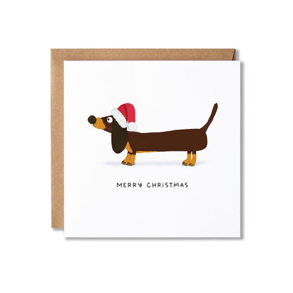 A square white Christmas greeting card with a brown dachshund wearing a red Santa hat, shown with a brown kraft envelope behind it on a light gray background.