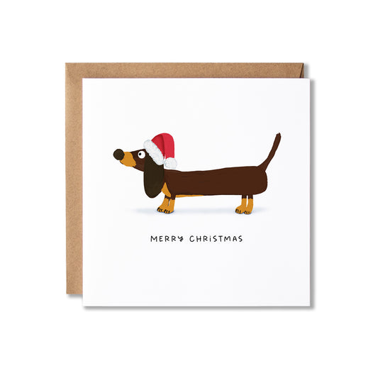 A square white Christmas greeting card with a brown dachshund wearing a red Santa hat, shown with a brown kraft envelope behind it on a light gray background.