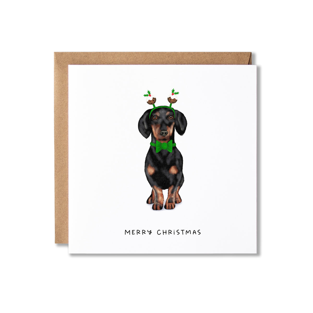 A square white Christmas greeting card with a brown kraft envelope behind it, featuring an illustration of a dachshund wearing a green bow tie and reindeer antlers, with the text 'MERRY CHRISTMAS' at the bottom.