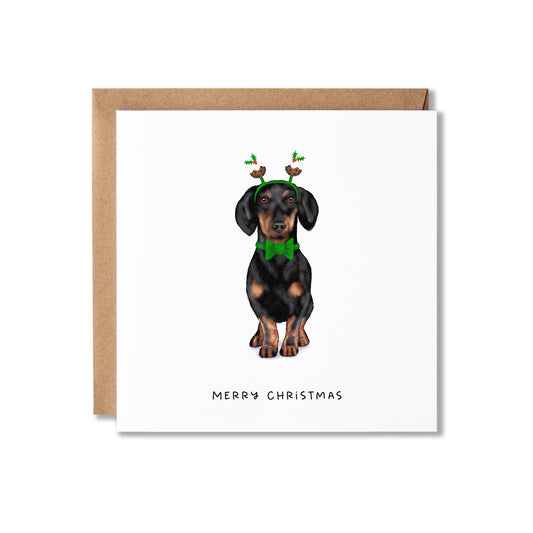 A square white Christmas greeting card with a brown kraft envelope behind it, featuring an illustration of a dachshund wearing a green bow tie and reindeer antlers, with the text 'MERRY CHRISTMAS' at the bottom.