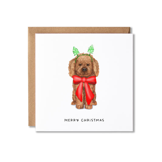 A square Christmas greeting card with a brown kraft envelope behind it. The card features a watercolor illustration of a brown dog wearing green leaf antlers and a large red bow, with the text 'MERRY CHRISTMAS' printed below.