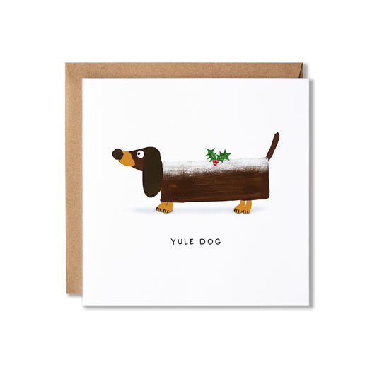 A square greeting card with a brown kraft envelope behind it, featuring a cartoon dachshund covered in snow with a small holly sprig and the text 'YULE DOG' below.