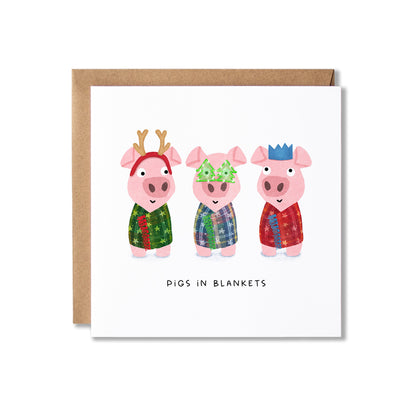 A square white greeting card with a brown kraft envelope behind it, featuring three cartoon pigs in colorful patterned blankets and the text 'PIGS IN BLANKETS' below.