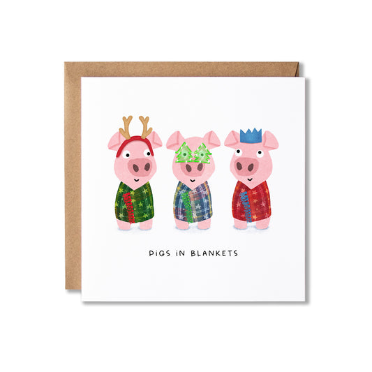 A square white greeting card with a brown kraft envelope behind it, featuring three cartoon pigs in colorful patterned blankets and the text 'PIGS IN BLANKETS' below.