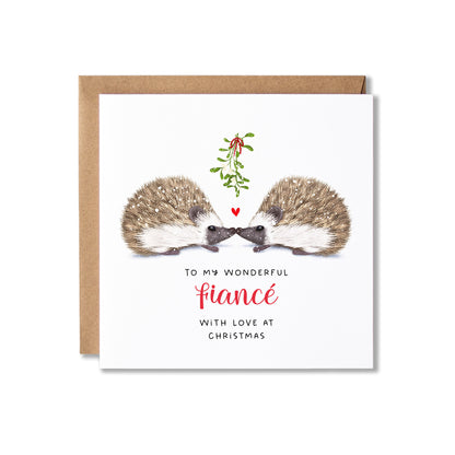 A square Christmas greeting card with a kraft envelope behind it. The card features an illustration of two hedgehogs facing each other with a small red heart and mistletoe between them, above the text 'TO MY WONDERFUL fiancé WITH LOVE AT CHRISTMAS' in black and red lettering on a white background.