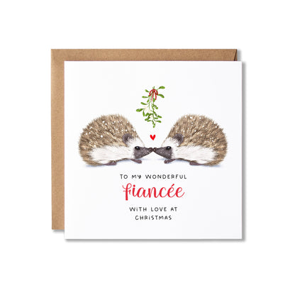 A square Christmas greeting card with a kraft envelope behind it, featuring an illustration of two hedgehogs facing each other under a sprig of mistletoe, with the text 'TO MY WONDERFUL fiancée WITH LOVE AT CHRISTMAS' on a white card background.