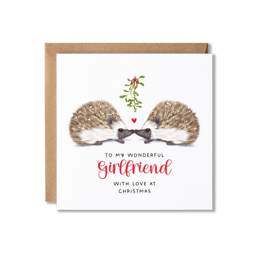 A square Christmas greeting card with a kraft envelope behind it. The card features an illustration of two hedgehogs facing each other under a small mistletoe sprig and red heart, with the text 'TO MY WONDERFUL Girlfriend WITH LOVE AT CHRISTMAS' in black and red lettering on a white background.