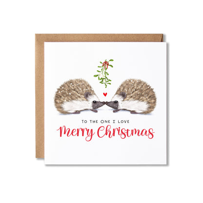 A square Christmas greeting card with a kraft envelope behind it, featuring an illustration of two hedgehogs facing each other under a small mistletoe sprig and red heart, with the text 'TO THE ONE I LOVE Merry Christmas'.
