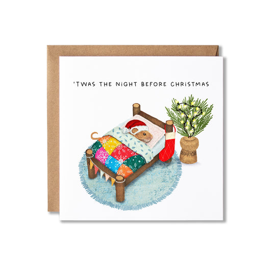 A square Christmas greeting card with a kraft envelope behind it, featuring an illustration of a small mouse wearing a Santa hat in a bed with a colorful quilt, a red stocking, and a potted plant. The card reads, "'Twas the night before Christmas."