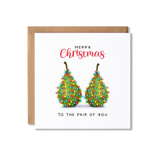 A square Christmas greeting card with a kraft envelope behind it. The card has a white background, red script text reading 'Merry Christmas', and an illustration of two green pears decorated with colourful string lights, with black text below reading 'To the pair of you'.