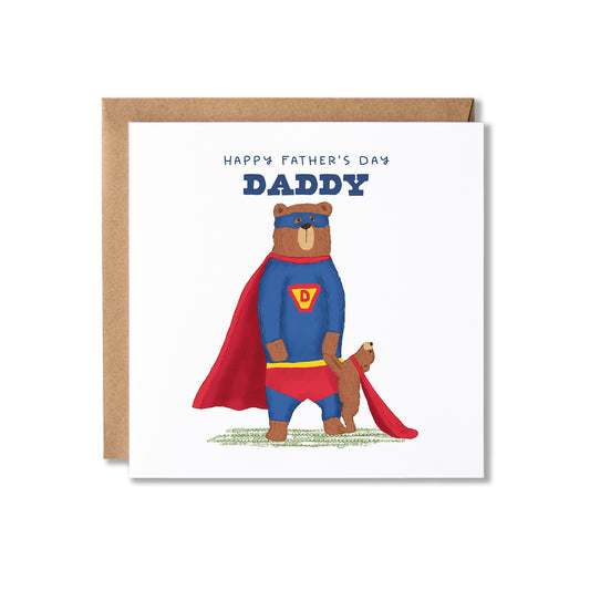 A square white Father's Day greeting card with a brown kraft envelope behind it. The card features an illustration of a bear dressed as Superman with a red cape and blue suit, holding hands with a bear cub, with the text 'HAPPY FATHER'S DAY DADDY' above.