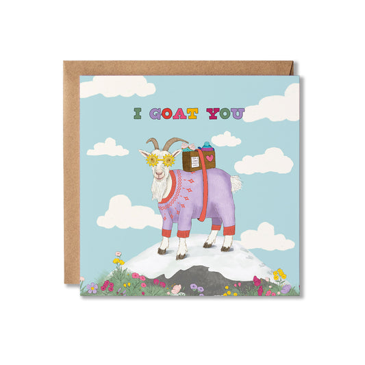 A square greeting card with a brown envelope behind it, featuring a whimsical illustration of a goat wearing a purple sweater carrying a basket of tea and cake, standing on a snowy peak with flowers and clouds against a light blue sky. The card reads 'I GOAT YOU' in multicoloured letters.