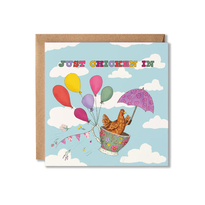 A square greeting card with a brown kraft envelope behind it, featuring a whimsical illustration of a brown chicken in a floral teacup with a purple umbrella and colourful balloons floating through the sky. The card reads 'JUST CHICKEN IN' in multicoloured letters against a light blue sky with white clouds.