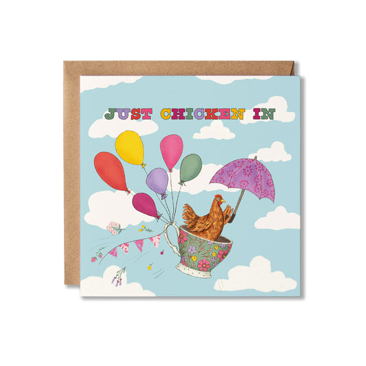 A square greeting card with a brown kraft envelope behind it, featuring a whimsical illustration of a brown chicken in a floral teacup with a purple umbrella and colourful balloons floating through the sky. The card reads 'JUST CHICKEN IN' in multicoloured letters against a light blue sky with white clouds.