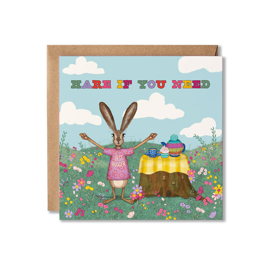 A square greeting card with a brown kraft envelope behind it, featuring an illustrated rabbit standing in a flower field with arms outstretched beside a small table. The card reads 'HARE IF YOU NEED' in multicolored letters against a light blue sky with clouds.